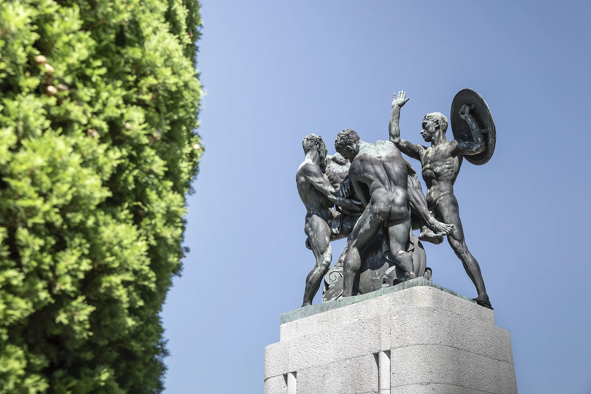 Monumento ai Caduti di Trieste con scultura bronzea su basamento in pietra, immerso nel verde e illuminato dalla luce naturale del cielo
