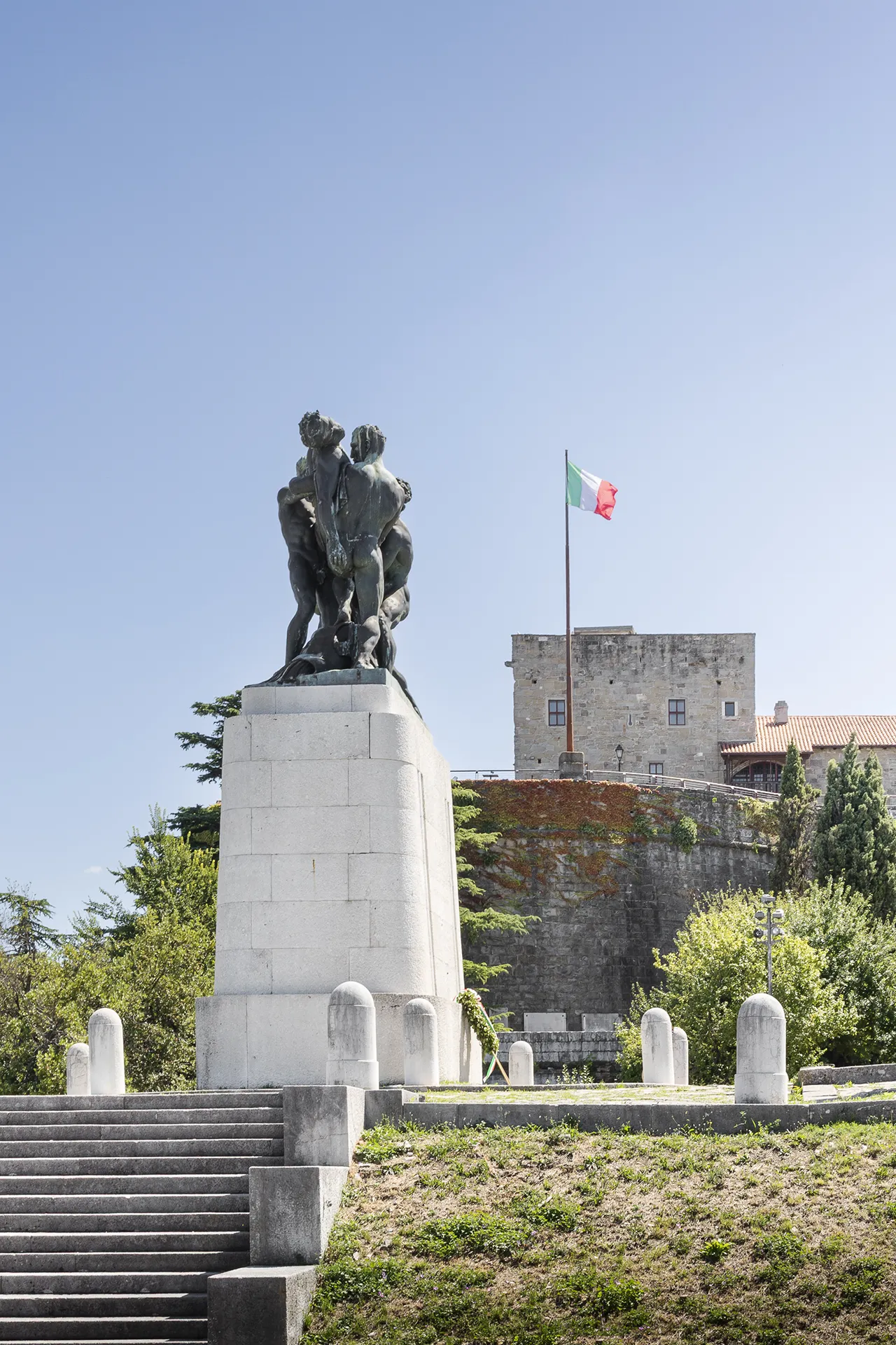 Monumento ai Caduti di Trieste con il Castello di San Giusto sullo sfondo, vista panoramica con scalinata in pietra, vegetazione e cielo sereno